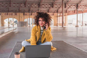 Positive young curly haired female freelancer in casual clothes smiling and on a phone call on mobile phone while drinking coffee during break from work on laptop on bench - ADSF44503