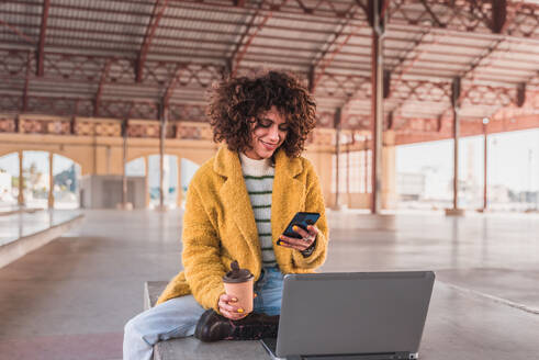 Positive young curly haired female freelancer in casual clothes smiling and surfing internet on smartphone while drinking coffee during break from work on laptop on bench - ADSF44504