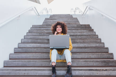 Full body of positive curly haired female freelancer in casual wear sitting on stairs and having phone call while working remotely on laptop and looking away on street - ADSF44505