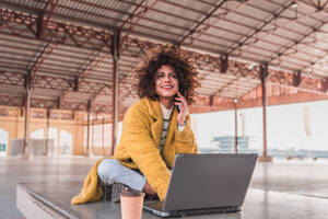 Positive young curly haired female freelancer in casual clothes smiling and on a phone call on smartphone while drinking coffee during break from work on laptop on bench - ADSF44506