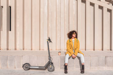 Full body of dreamy young curly haired female in casual clothes and sunglasses sitting on ledge with cup of takeaway coffee near electric scooter with eyes closed on street - ADSF44509