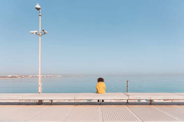 Back view of distant female in yellow coat sitting on bench and admiring picturesque seascape from embankment against cloudless blue sky - ADSF44514