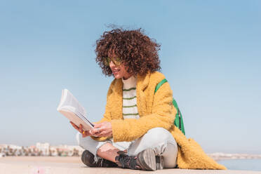 Full body of calm curly haired female in casual clothes and glasses sitting on border in lotus pose while reading interesting book against rippling sea - ADSF44515