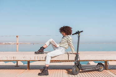 Full body side view of dreamy curly haired female in casual outfit and sunglasses sitting reading a book on embankment near electric scooter against sea - ADSF44516