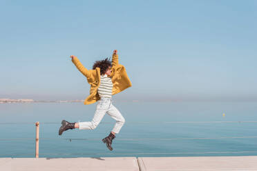 Full body of carefree young female in casual outfit jumping above ground and having fun with raised arms on embankment against sea - ADSF44518