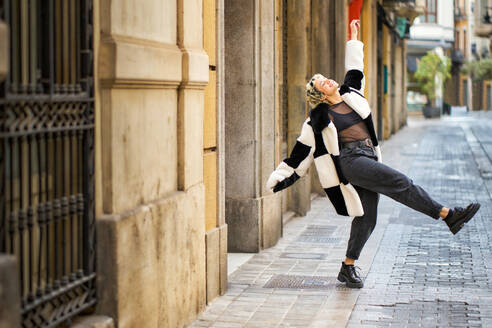 Full body of excited young female in stylish outfit raising arms and leg while celebrating achievement on paved street - ADSF44526