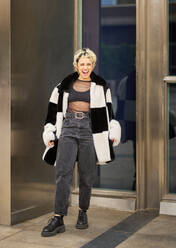 Full body of excited young female in casual outfit and fashionable fur coat standing on paved street with opened mouth and looking at camera against modern building - ADSF44535