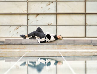 Positive young female in stylish fur coat lying on concrete border near glass floor and looking at camera on street - ADSF44538