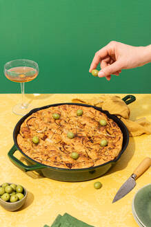 High angle of anonymous person garnishing olives on flat oven baked focaccia bread in pan while serving with wine in glass on yellow surface against green backdrop - ADSF44553