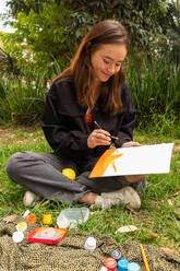 Happy young female artist sitting on grass and painting in paper with brush while resting at daytime against green plants - ADSF44558