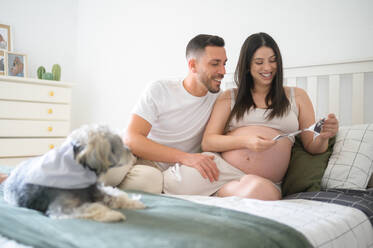 Cheerful young man with pregnant woman smiling and looking at ultrasound photo of baby while sitting on bed near dog in cozy bedroom - ADSF44577