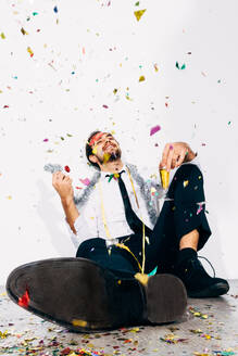Content male in formal wear with glass of alcoholic drink sitting on floor with confetti and looking up during Christmas holiday - ADSF44578