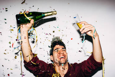 Happy male with glass and bottle of alcoholic drink under confetti looking up during party - ADSF44580