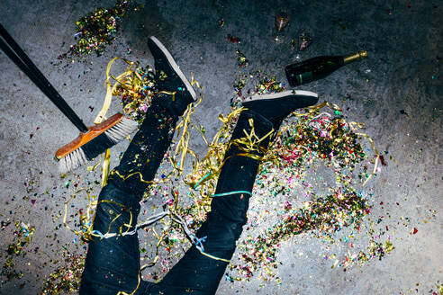 Top view of crop anonymous male lying on floor with colored paper streamers and bottle of champagne during festive occasion - ADSF44581