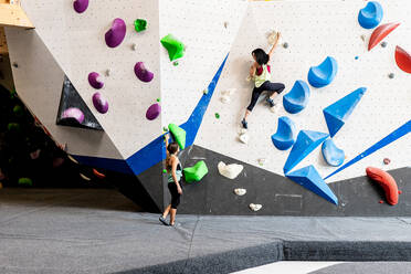 Back view of unrecognizable female alpinist in sportswear climbing bouldering wall with bright grips while anonymous friend standing on floor and guiding during practice in gym - ADSF44582