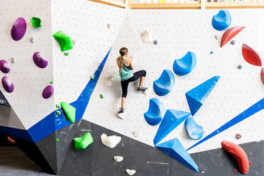 From below back view of unrecognizable female athlete in sportswear and climbing shoes grabbing grips while exercising on climbing wall in bouldering studio - ADSF44583