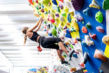 From below side view of anonymous female with shoes holding on to holds while climbing indoor bouldering wall with colorful grips during workout in modern gym - ADSF44586