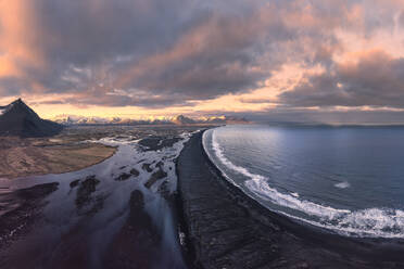 Aerial view of rocky mountains covered with snow located near sea against cloudy sky during sunset in Iceland - ADSF44598