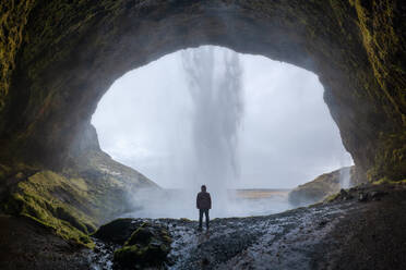 Back view of unrecognizable tourist with backpack standing on edge of rocky cliff and admiring picturesque scenery of Seljalandsfoss waterfall - ADSF44601