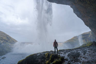 Back view of unrecognizable tourist with backpack standing on edge of rocky cliff and admiring picturesque scenery of Seljalandsfoss waterfall - ADSF44602