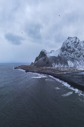 Drone view of sea waves washing stony beach with snowy mountains against cloudy sky in Iceland - ADSF44609