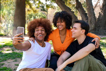 Happy multiracial friends in casual clothes smiling and taking self portrait on smartphone while spending time together in Ciutadella Park - ADSF44611