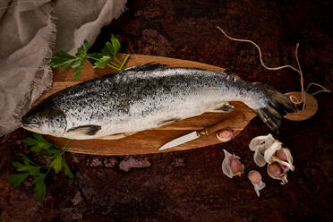 From above of uncooked fish with green leaves and knife placed on wooden cutting board near garlic - ADSF44614