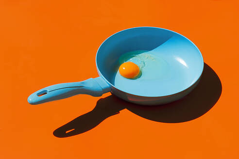 Whole raw egg yolk placed in blue frying pan with handle isolated against orange background - ADSF44637