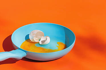 Blue frying pan with raw egg yolk and eggshell isolated on orange background - ADSF44638