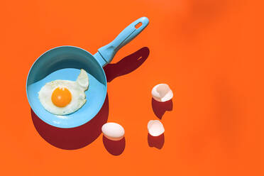 Top view of fried egg on blue frying pan near raw whole egg and eggshells with shadow isolated on orange background - ADSF44639