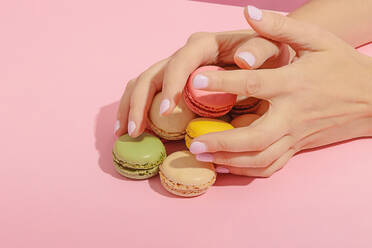 Unrecognizable female with manicure taking pile of tasty sweet multicolored macaroons from pink surface - ADSF44640