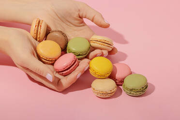 Unrecognizable female with manicure taking pile of tasty sweet multicolored macaroons from pink surface - ADSF44642