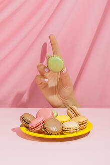 Unrecognizable female demonstrating delicious green macaroon near round plate with colorful macaroons on pink background in studio - ADSF44643