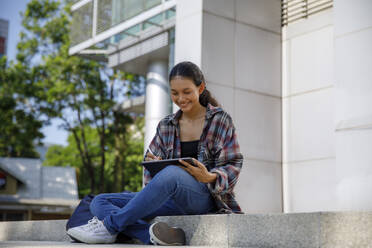 Lächelnder Student mit Tablet-PC im Schneidersitz vor der Universität - IKF00971