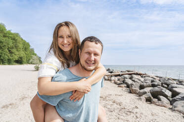 Smiling man giving piggyback ride to woman at beach - IHF01470