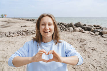 Smiling woman making heart sign at beach - IHF01475