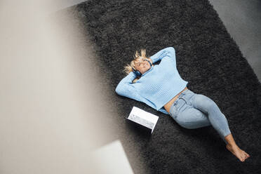 Happy freelancer wearing wireless headphones lying on carpet at home - JOSEF19863