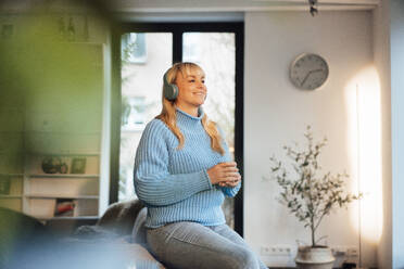 Happy woman wearing wireless headphones sitting on sofa at home - JOSEF19925