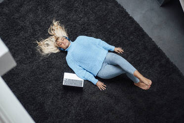 Freelancer wearing wireless headphones lying with eyes closed and laptop on carpet - JOSEF19938