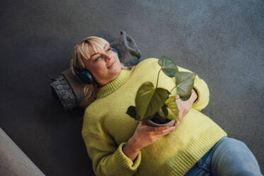 Lächelnde Frau mit drahtlosen Kopfhörern, die mit einer Topfpflanze auf dem Boden liegt - JOSEF19957