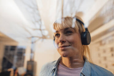 Smiling blond woman wearing wireless headphones seen through glass - JOSEF19967