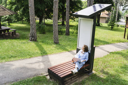 Woman charging laptop through solar charging point sitting at park - EKGF00256