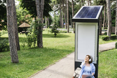 Lächelnde Frau, die in der Nähe einer Solarladestation im Park sitzt und Musik hört - EKGF00258