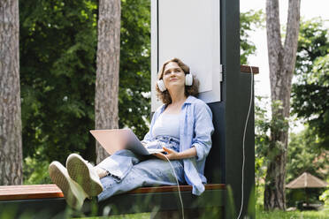 Lächelnde Frau, die mit ihrem Laptop an einer Solarladestation im Park sitzt und träumt - EKGF00261