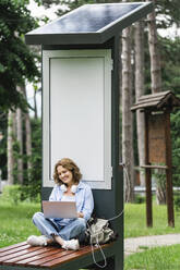Lächelnde Frau mit Laptop auf einer Bank in der Nähe einer Solarladestation im Park - EKGF00269