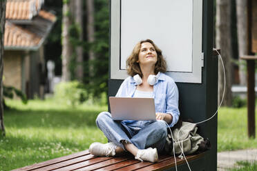 Thoughtful woman with laptop sitting near solar charging point at park - EKGF00270