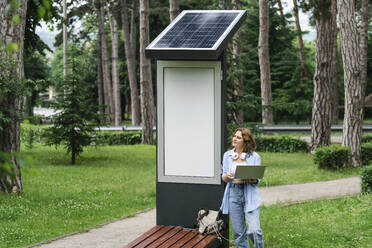 Frau mit Laptop steht in der Nähe einer Solarladestation im Park - EKGF00271