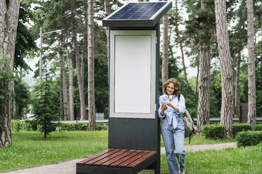 Frau mit Mobiltelefon in der Nähe einer Solarladestation im Park - EKGF00273