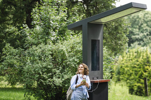 Smiling woman standing near solar charging point at park - EKGF00276