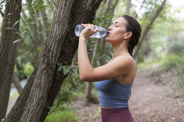 Woman drinking water near tree at forest - LMCF00296